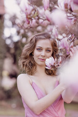 Fototapeta premium Spring flowering. Portrait of a young blonde girl in the branches of a blooming magnolia. Open aperture with light blurring and illumination.