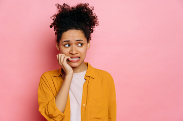 Fototapeta na wymiar Stunned african american young woman, in casual wear, looking stressed and nervous with hands on mouth biting nails, looking aside, is going through, stands on isolated pink background
