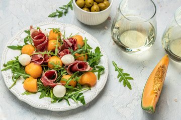 mediterranean salad with prosciutto or jamon, mozzarella and green basil leaves and Cantaloupe melon on white table, traditional Spanish and Italian appetizer served with wholemeal grissini, top view