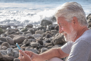Relaxed and smiling adult bearded senior man sitting on the rock beach using mobile phone. White-haired elderly grandfather enjoying technology and social and free time during retirement