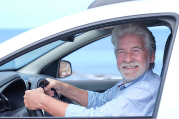 Beautiful adult active white-haired senior man sitting inside the car touching the steering wheel looking at camera smiling. Elderly grandparent with white beard enjoying travel. Horizon over water