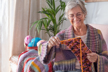 Senior smiling woman sitting on sofa at home while knitting and looking at her work in progress....