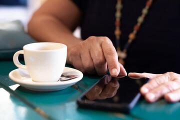 Fototapeta na wymiar Addicted adult mature caucasian woman sitting at the table with white cup of coffee while texting with mobile phone. Unrecognizable female people enjoying social and technology using smartphone