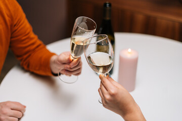 Close-up top view of unrecognizable happy couple clinking glasses of wine at table with candles in dark room. Loving man and woman celebrating anniversary, Valentines day, enjoying romantic date.