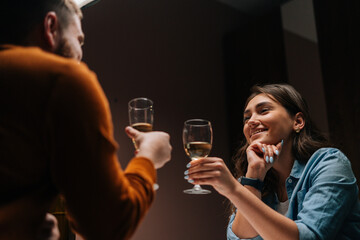 Low-angle view to smiling pretty young woman talking celebration toast and clinking white wine glass with husband during romantic dinner. Love couple celebrating anniversary or Valentines day.