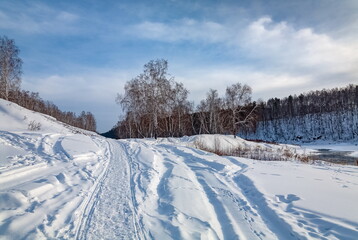 Winter landscape with birch trees on the mountain bank of the river, snow and blue sky with clouds