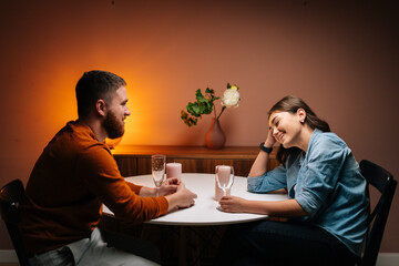 Side view of happy young couple enjoying talking, having fun together celebrating Valentines day dining during romantic dinner date, sitting together at table with candles in cozy dark living room.
