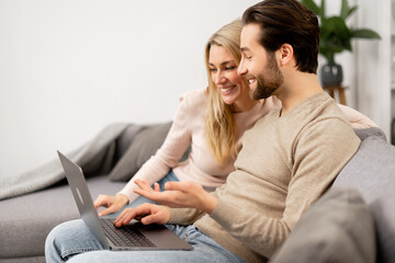 Happy girlfriend and her boyfriend looking at the laptop screen while sitting on the sofa, watching movies. Caucasian couple in love spending leisure time with laptop at home