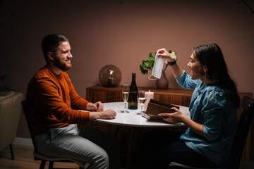 Funny couple husband and wife celebrating anniversary at home sitting at table. Cheerful young woman opening box and takes out medical protective mask. Pretty female receives red gift box with present