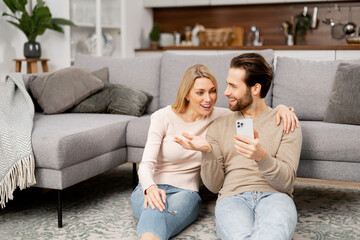 Excited and overjoyed caucasian young couple sitting near the couch at home and holding smartphone. Man and woman cant believe at something, celebrating good news or win, received money on account
