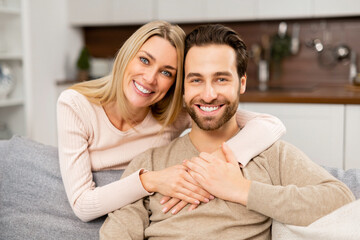 Fototapeta premium Portrait of lovely couple at home. Handsome man sitting on the comfortable couch, beautiful blonde woman hugging his from the back. Spouses spend time together