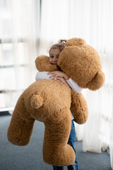 A little girl plays with a big teddy bear. She is dressed in a white jumper. Childhood. Lifestile.