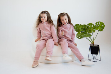 Two cute little girls in a pink overalls sits in a white chair. White background. Fashion....