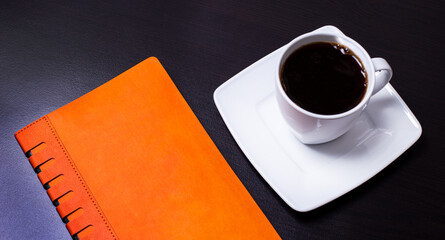 On a black background, a white cup of coffee and an orange diary. Business concept