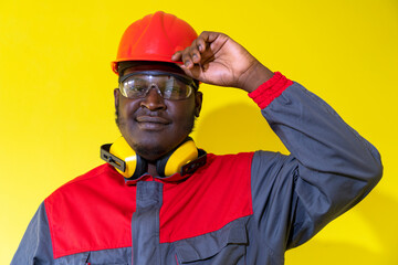 African American Worker In Protective Workwear Against Yellow Background. Portrait Of Black Industrial Worker In Red Helmet, Protective Eyewear, Hearing Protection Equipment And Work Uniform.