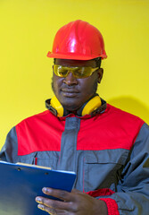 African American Worker In Personal Protective Equipment Writing On Clipboard Against Yellow Background. Waist Up Portrait Of Black Industrial Worker In Red Helmet, Safety Goggles And Work Uniform.