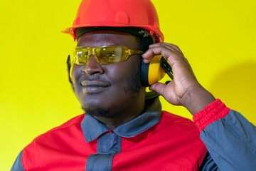 Young African-American Worker In Personal Protective Equipment Against Yellow Background. Portrait Of Black Industrial Worker In Red Helmet, Hearing Protection Equipment And Work Uniform.