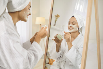 Happy young Caucasian woman look in mirror do facial clay moisturizing mask. Smiling girl after shower do morning beauty face procedures, take care of skin. Skincare and cosmetology concept.