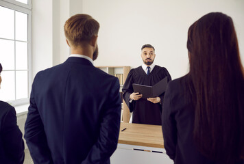 Lawyer, defendant and audience standing in courtroom and listening to judge giving judgment. Young male judge in gown enforces punishment on accused man. Back rear view. Law, justice, trial concept