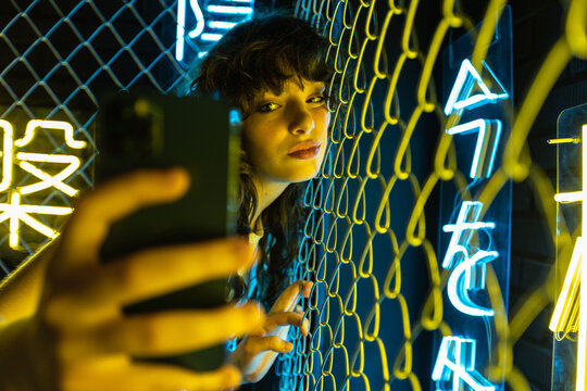 A young woman with curly hair takes a selfie near a yellow metal grid and in neon lighting.