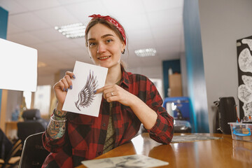 Portrait of smiling artist woman sitting at table in tattoo salon with drawing in her hands