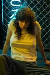 A young woman with curly hair in a white T-shirt sits near a yellow metal mesh.