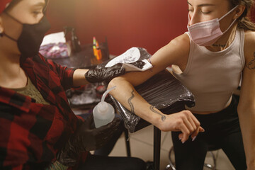 Portrait of female master and client wearing mask sitting in tattoo salon, tattoo master wiping...