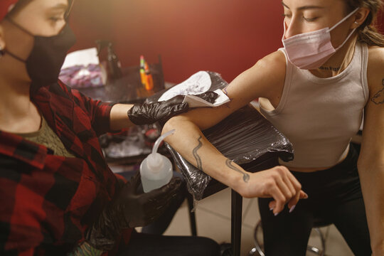 Portrait of female master and client wearing mask sitting in tattoo salon, tattoo master wiping client hand with napkin