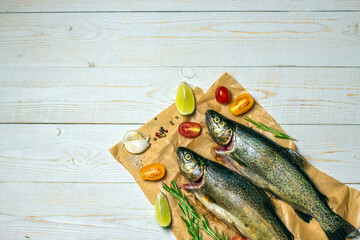 Raw fish, trout with on craft paper with ingredients for cooking. Fish, pepper, garlics, tomatoes, lemon and rosemary on white wooden table. Healthy food and dieting concept flat lay, with copy space.