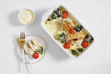Baked chicken fillet with broccoli and tomatoes in a white form, served with plate and piece of it on a white background, keto diet, top view