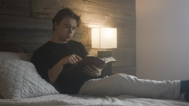Young white male doing his own bible study and devotionals. He is in his bedroom relaxing, reading scripture and questioning his reading.