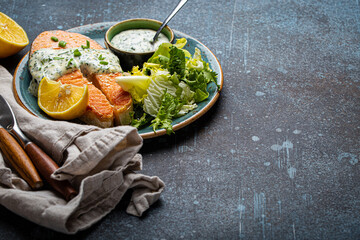 Healthy food meal cooked grilled salmon steak with white dill sauce and green salad leafs on plate on rustic concrete stone background table angle view, diet healthy nutrition dinner copy space