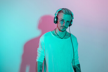 Fashion man model hipster with glasses listen to music in headphones in studio with pink blue light