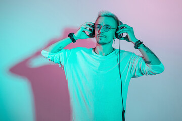 Handsome hipster man in fashionable clothes with glasses puts on headphones and listens to music in studio with colored creative light.