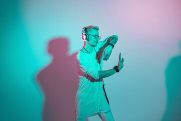 Stylish handsome man with headphones and glasses listening to music and dancing in the studio with creative pink and blue light. Guy to the rhythm of the dance