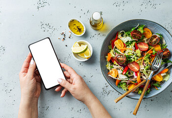 Smartphone in woman’s hands next to vegetable salad. Using phone while eating lunch.