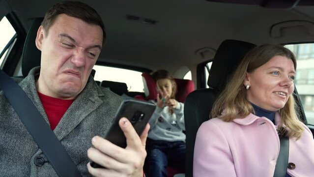 unhappy married couple with their daughter on trip. woman is driving car with disgusted and depressed person sitting in passenger seat looking at smartphone screen, he needs to pay his debts immediate