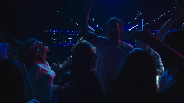 Group of people dancing with hands above heads around DJ in club. Video effects in background. Nightlife, modern music and entertainment concept.