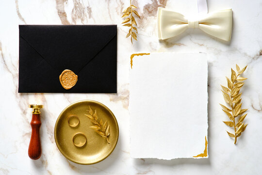 Elegant wedding stationery set. Wedding invitation card template, black wedding envelope with wax seal stamp, golden ring, golden floral branches, bow tie on marble table. Flat lay, top view.
