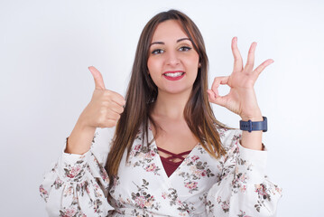 young Arab woman wearing floral dress over white background  smiling and looking happy, carefree and positive, gesturing victory or peace with one hand