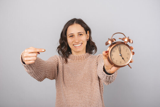 Smiling woman is pointing at the alarm clock she is holding. It is five o clock. Portrait over grey background.