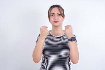 Obraz na płótnie Canvas Displeased annoyed young Arab woman wearing striped t-shirt over white wall clenches fists, gestures pissed, ready to revenge, looks with aggression at camera stands full of hate, being pressured