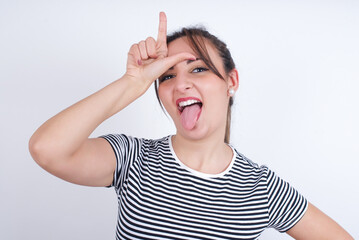 Funny young Arab woman wearing striped t-shirt over white background makes loser gesture mocking at someone sticks out tongue making grimace face.