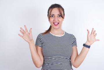 Delighted positive young Arab woman wearing striped t-shirt over white background  opens mouth  and arms palms up after having great result