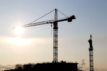 Silhouettes of construction cranes and unfinished residential building on sunshine background. Housing construction, apartment block in city