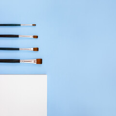 Paint brushes and a watercolor sketchbook on a blue background. Minimal concept of freedom of creativity and self-expression. Square orientation, copy space