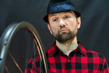 A fashionable bicycle mechanic in a red shirt and a black hat holds a wheel in his hand. Assembling wheels from a carbon rim using professional equipment for tension spokes. Bicycle repair.