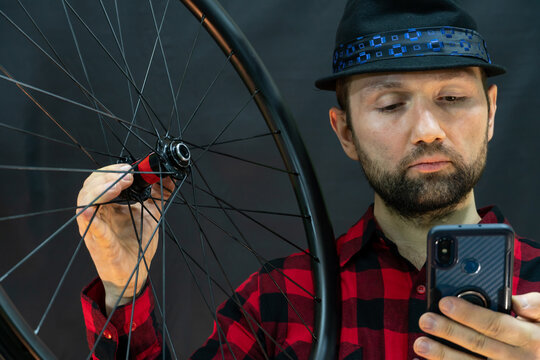 A fashionable bicycle mechanic in a red shirt and a black hat holds a wheel and a mobile phone in his hand. Assembling wheels from a carbon rim using professional equipment for tensioning spokes.