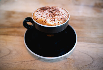 Cappuccino froth with chocolate sprinkles in a black cup and saucer with with white rim. The drink is on a wooden table.