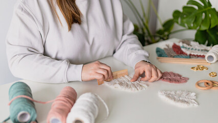 Woman working out last details of an almost-finished macrame piece, combing ropes inside a home room. Close up. Natural cotton threads and wooden beads. Female hobby. Wall hanging decor.
